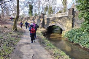 4 walkers with backpacks walking up a path next to a river with a bridge in the background. It looks wintery and cold.