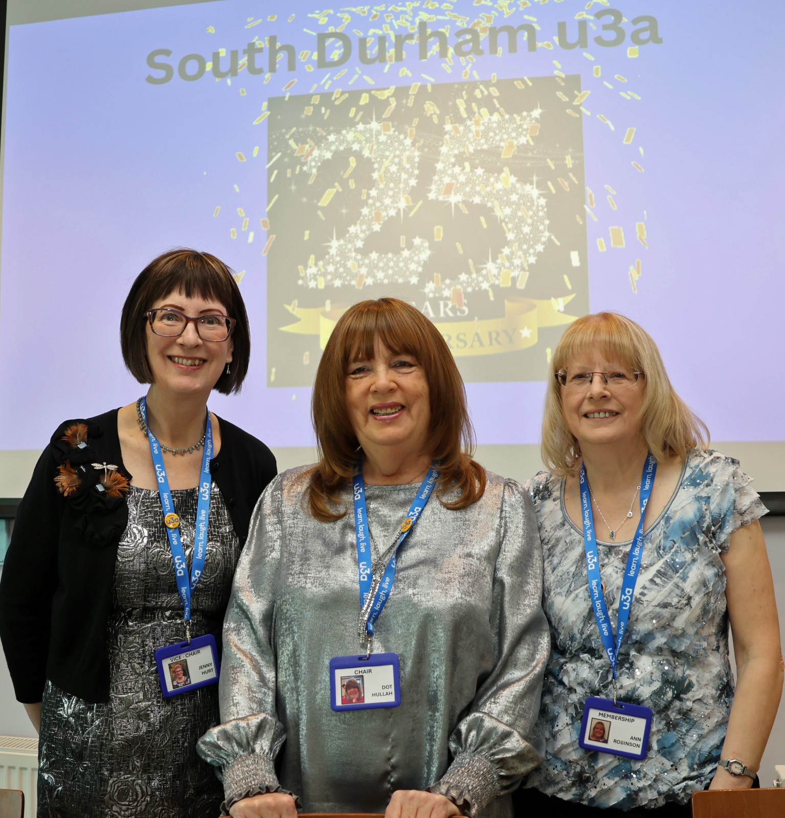 3 u3a members standing in front of a slideshow marking the 25th anniversary of South Durham u3a