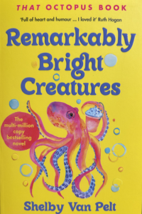 The book cover of Remarkably Bright Creatures. The cover is bright yellow with a drawn pink octopus on the front, holding a cupcake in its hand. At the top of the book it reads, 'That Octopus Book', below is an author quote, 'Full of heart and humour... I loved it' Ruth Hagan' and below that, the title for the book 'Remarkably Bright Creatures'.