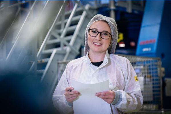 A woman in a white apron and white net covering her hair is holding pieces of paper and standing in a space station