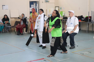 Two women batting, a man behind wearing a white u3a vest is sitting down 