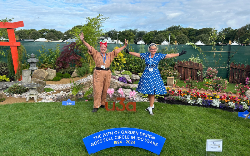 Two women are dressed in clothes inspired by vintage fashion - the woman on the left is wearing a beige boiler suit with a red bandana tied round her head, in the style of Rosie the Riveter. The woman to the left of her is wearing a bright blue 50s inspired dress. They are standing on the grass of a show garden. Behind them are various flower beds - a colourful 1950s flower bed, a bridge leading to a Japanese styled section of the garden. They are standing next to a wooden sign that reads u3a, and in front of them, a blue plaque that says 'The path of garden design goes full circle in 100 years.'