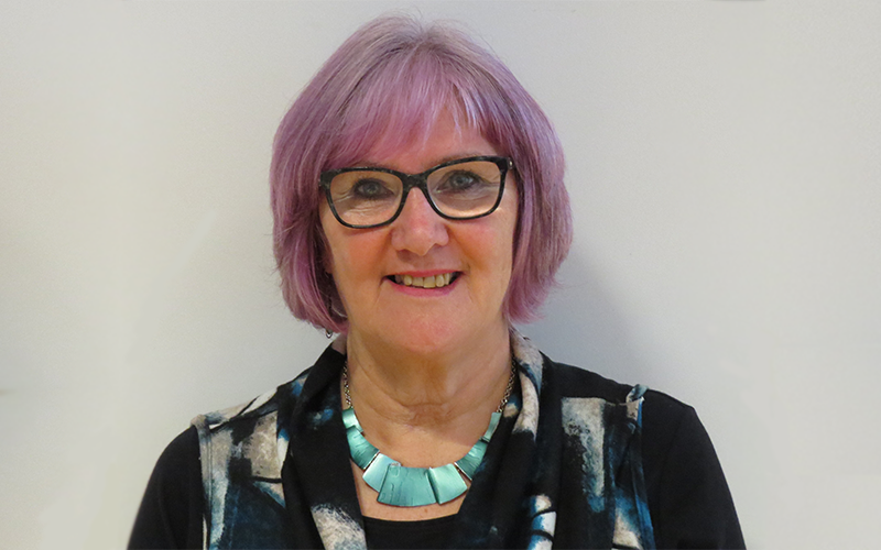A woman with short pink hair is standing against a grey background and smiling.