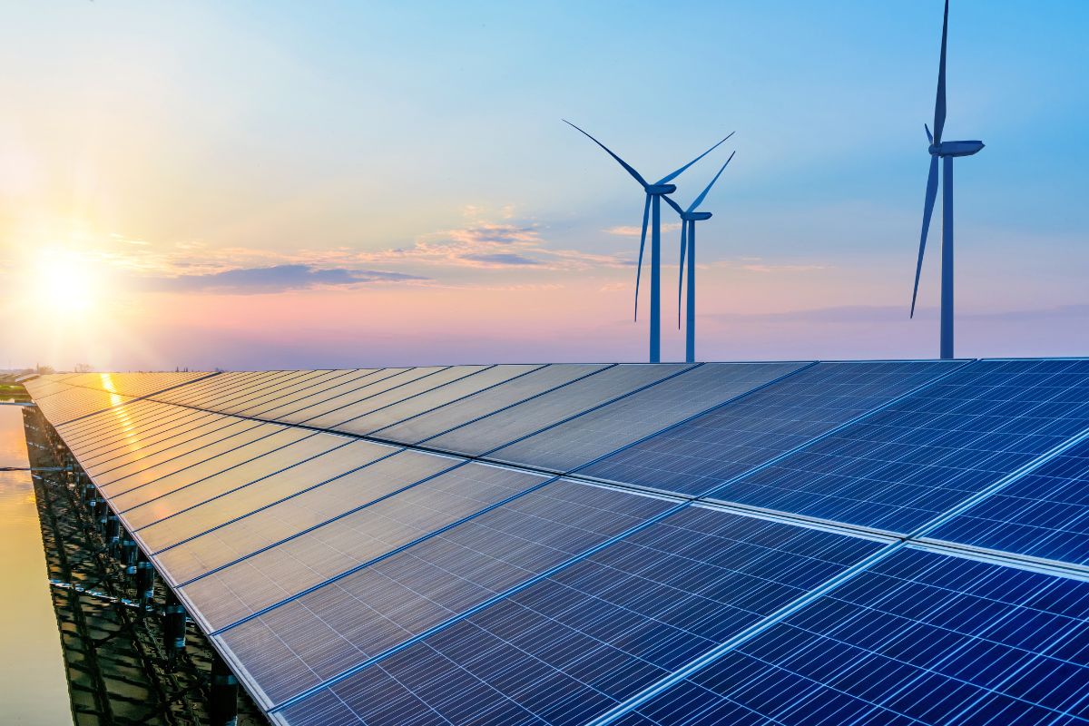 Rows of solar panels, with wind turbines in the background, are set against a blue and pink sky
