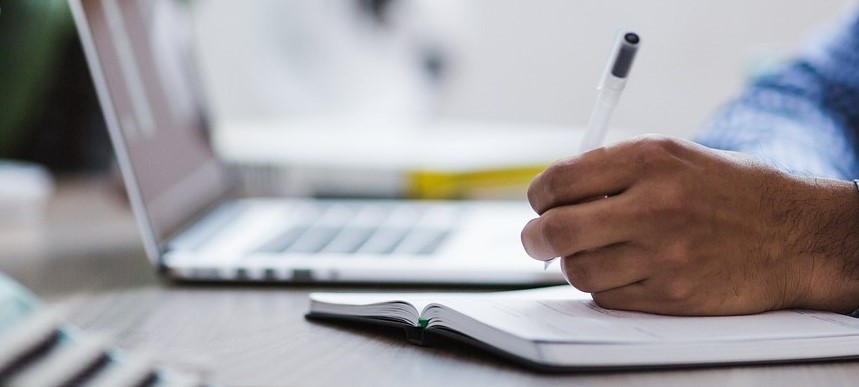 hand holding pen writing next to laptop