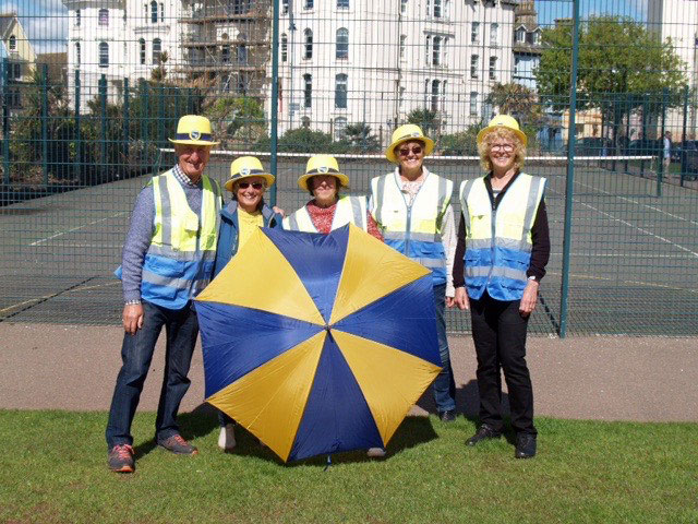five people in blue and yellow high vis jackets smiling at camera holding blue and yellow umbrella 