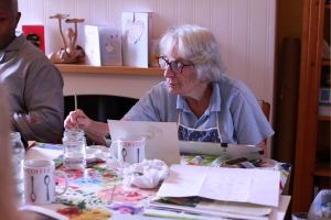 Marion painting; she is inside a house, and the table is filled with painting supplies.