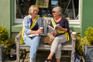 Two women reading books on a bench in summer, wearing u3a sashes