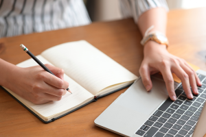 A woman's hands - one hand is writing notes in a notebook, the other is typing on a laptop.