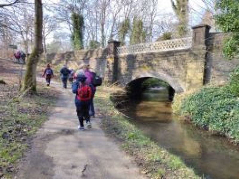 a photo of walkers on a path by a river with a bridge on the right
