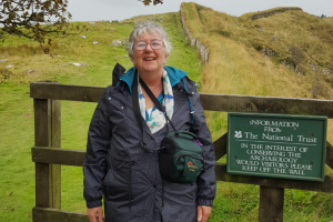 a woman with short grey hair smiling next to a national trust sign on the hadrian's wall path. Green fields behind her and she wears a blue raincoat.