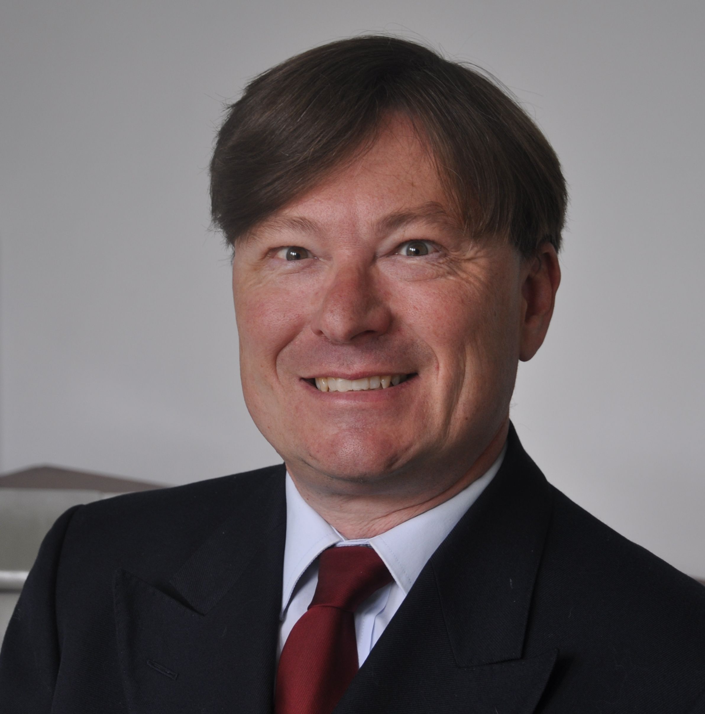 A man in a suit jacket and white shirt, and red tie. He has brown hair.