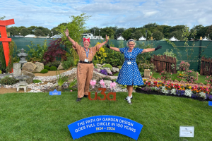 Two women are dressed in clothes inspired by vintage fashion - the woman on the left is wearing a beige boiler suit with a red bandana tied round her head, in the style of Rosie the Riveter. The woman to the left of her is wearing a bright blue 50s inspired dress. They are standing on the grass of a show garden. Behind them are various flower beds - a colourful 1950s flower bed, a bridge leading to a Japanese styled section of the garden. They are standing next to a wooden sign that reads u3a, and in front of them, a blue plaque that says 'The path of garden design goes full circle in 100 years.'
