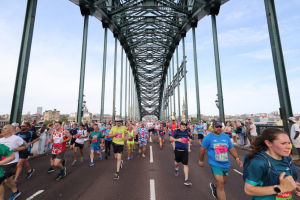Runners in colourful running gear run towards the camera across a tall iron bridge with blue skies in the background