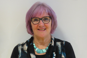 A woman with short pink hair is standing against a grey background and smiling.